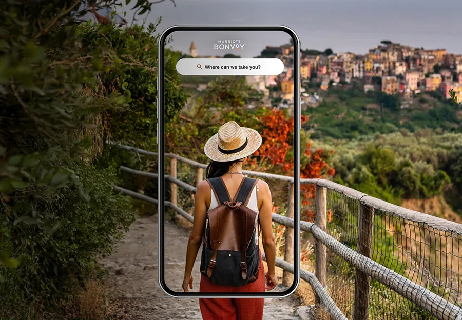A traveler with a hat and backpack walking a scenic trail, shown through a smartphone frame displaying the Marriott Bonvoy search bar.